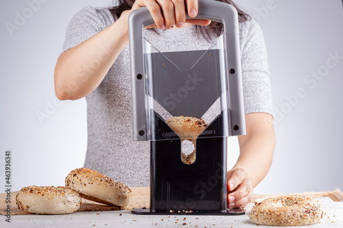 Cacuasian woman wearing short sleeve t shirt is slicing fresh baked bagels using manual bagel slicer. She places the bagel inside the holder and puts sharp blade through it using the handle. photo