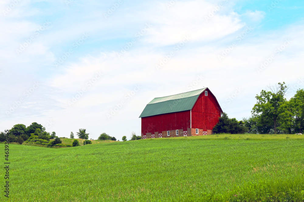 bright red farm barn building on a bright green pasture hillside under cloudy blue sky