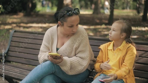 Mom and daughter are talking on a park bench. The child shows new trends on the Internet.