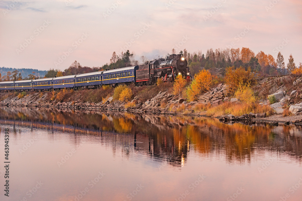 Retro steam train moves along the lake at autumn sunset time.