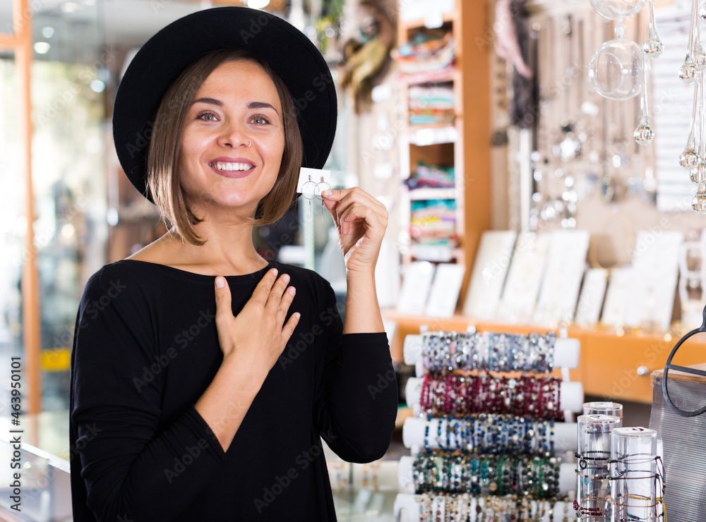 Adult woman customer in hat trying earrings in the shop of bijouterie.