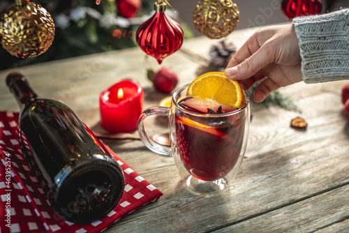 Woman in a sweater is holding a clear mug with fragrant spicy mulled wine decorated with fruits and spices. Concept of a cozy festive atmosphere, New Year and Christmas mood