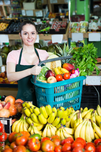 Smiling attractive saleswoman with shopping basket with greengrocery in grocery
