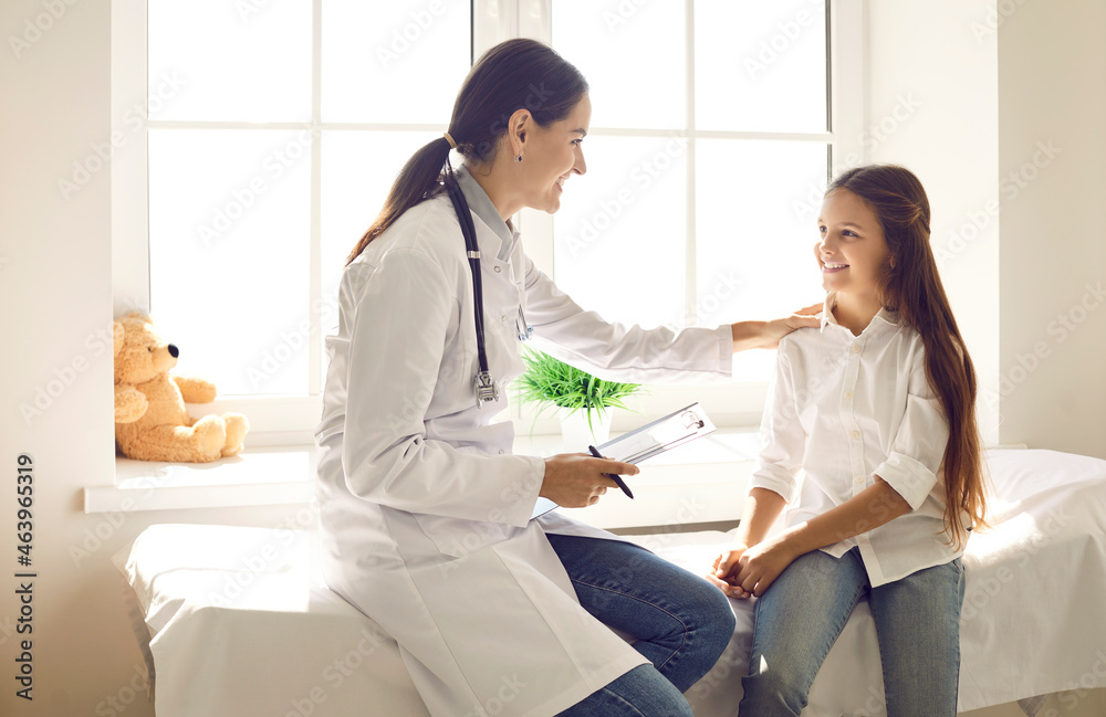 Friendly doctor talking to child and touching her shoulder to support her at medical checkup. Happy teen girl sitting on examination couch in exam room and listening to female pediatrician's advice - obrazy, fototapety, plakaty 