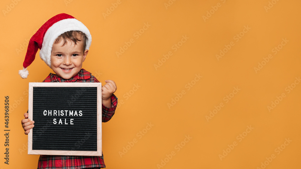 A smiling boy in a red Santa hat and pajamas, with a board with white letters and the inscription Merry Christmas in his hands. A place for your text and advertising. A smiling boy in a red Santa hat and pajamas, with a board with white letters and the inscription Merry Christmas in his hands. A place for your text and advertising.