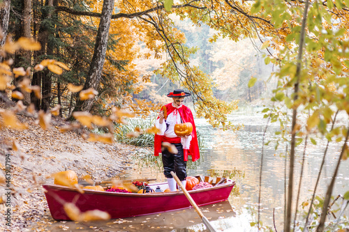 young man dressed as gondolier in gondola boat decorated with pumpkins on pond in autumn park celebrates Halloween and having fun, concept of Halloween carnival or costume party