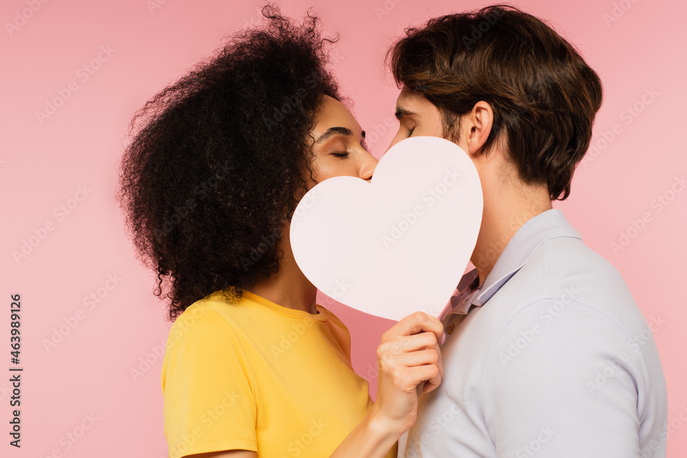 interracial couple with closed eyes kissing behind blank paper heart isolated on pink. interracial couple with closed eyes kissing behind blank paper heart isolated on pink.
