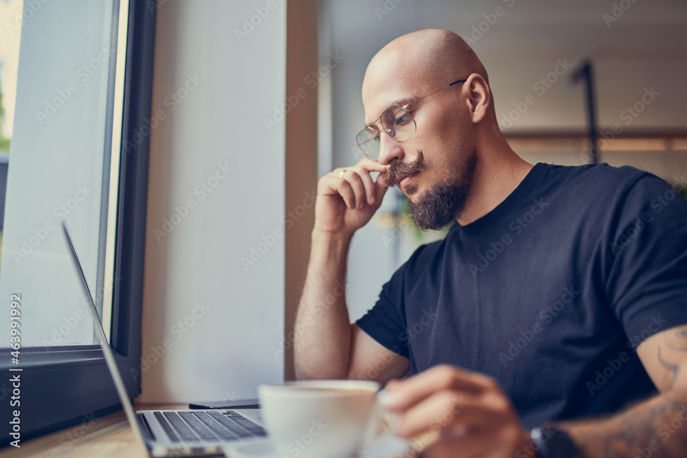 Focused hipster millennial man works at laptop while sitting in cafe with coffee. Freelance concept