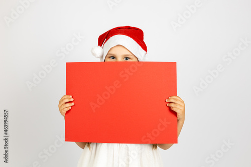 portrait of happy girl with Santa Claus hat hiding behind red empty poster in hands, isolated on white background. 