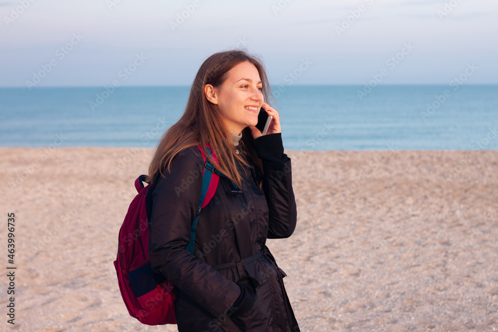 Happy young woman in black raincoat empty autumn sea beach. Smiling girl drinking tea with thermo can bottle. Lifestyle Happy young woman in black raincoat empty autumn sea beach. Smiling girl drinking tea with thermo can bottle. Lifestyle
