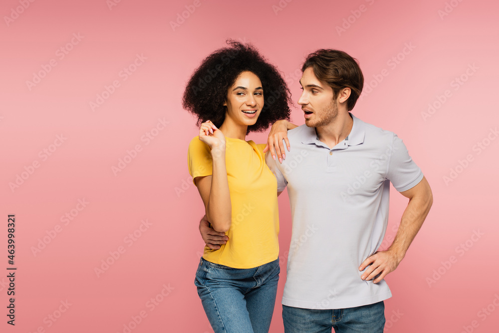 surprised man hugging cheerful hispanic woman while standing with hand on hip isolated on pink. surprised man hugging cheerful hispanic woman while standing with hand on hip isolated on pink.