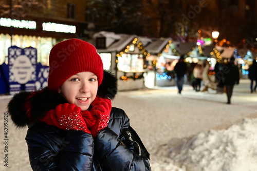 pretty smiling girl in warm clothes, wearing red hat, scarf and gloves stands against background of winter decorated street on New Year's Eve. Concept of Christmas fairs and sales