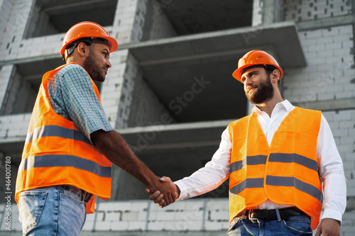 Two men engineers in workwear shaking hands against construction site.