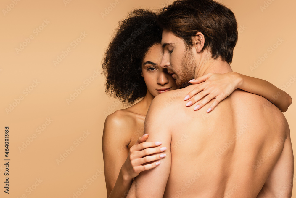 Naklejka premium seductive latin woman looking at camera while hugging sexy shirtless man isolated on beige.