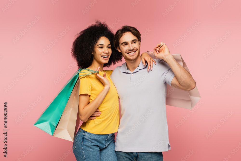 cheerful interracial couple embracing while holding shopping bags and looking away isolated on pink. cheerful interracial couple embracing while holding shopping bags and looking away isolated on pink.