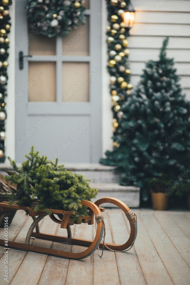 Fototapeta premium Door of wooden house decorated with festive lights and Chrismas wreath. Sled with pine. Holiday house decoration.