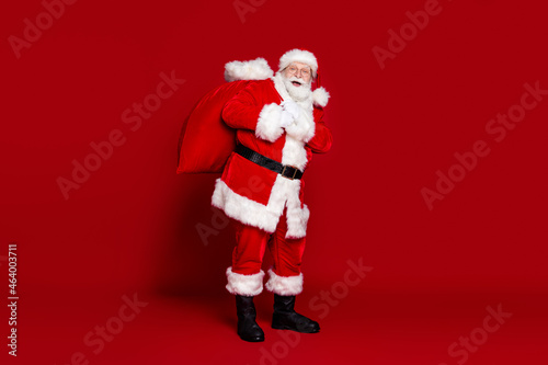 Photo of funny excited man pensioner dressed santa claus costume carrying big christmas sack smiling isolated red color background