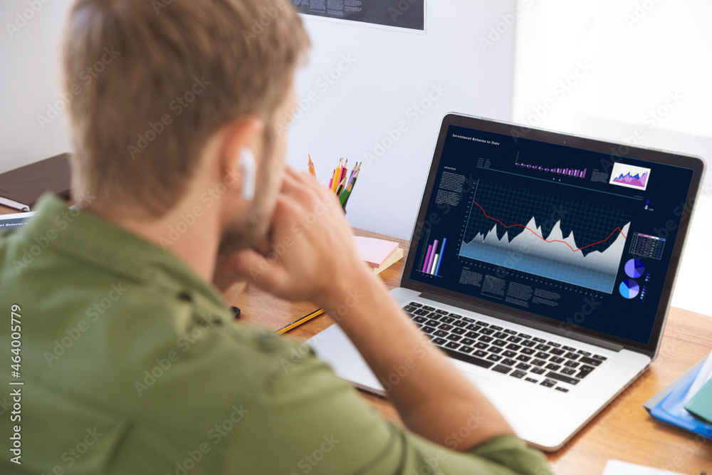 Caucasian businessman sitting at desk, using laptop with statistical data on screen Caucasian businessman sitting at desk, using laptop with statistical data on screen