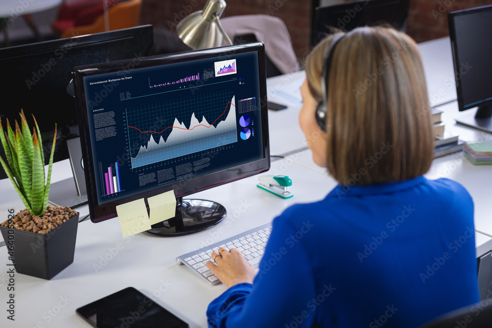 Caucasian businesswoman sitting at desk, using computer with statistical data on screen Caucasian businesswoman sitting at desk, using computer with statistical data on screen