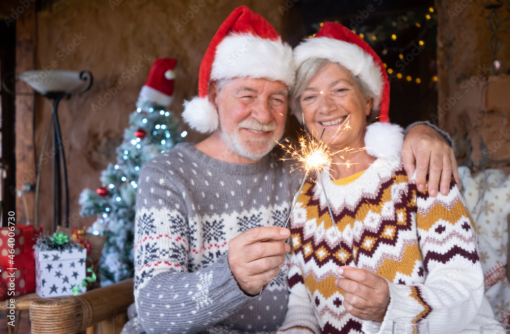 Senior smiling couple in Santa hats hugging at home at Christmas time with sparklers. Christmas tree, presents and decorations on background Senior smiling couple in Santa hats hugging at home at Christmas time with sparklers. Christmas tree, presents and decorations on background