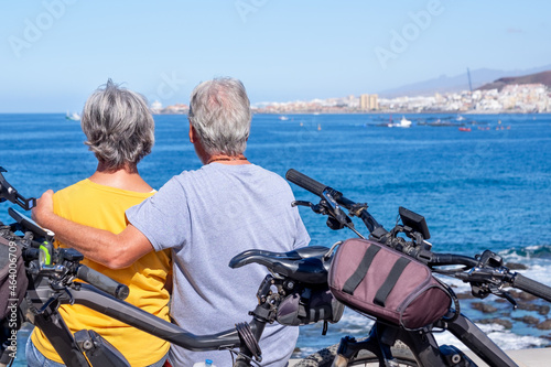 Rear view of senior couple sitting on the cliff in sea excursion with their bicycles, looking at the horizon. Active retirees enjoying healthy lifestyle