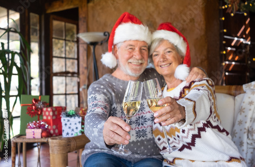 Defocused senior couple in Santa hats hugging at home at Christmas time toasting with wine glasses. Sitting inside a rustic mountain chalet with Christmas tree, presents and lights on background