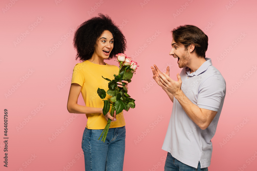 happy latin woman holding bouquet of roses near amazed man showing wow gesture isolated on pink. happy latin woman holding bouquet of roses near amazed man showing wow gesture isolated on pink.