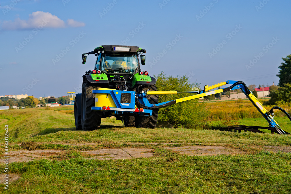 Traktor, z kosiarką na wysięgniku, koszący trawę na wale rzecznym . Tractor, with a mower on a boom, muting grass on the river shaft. - obrazy, fototapety, plakaty 
