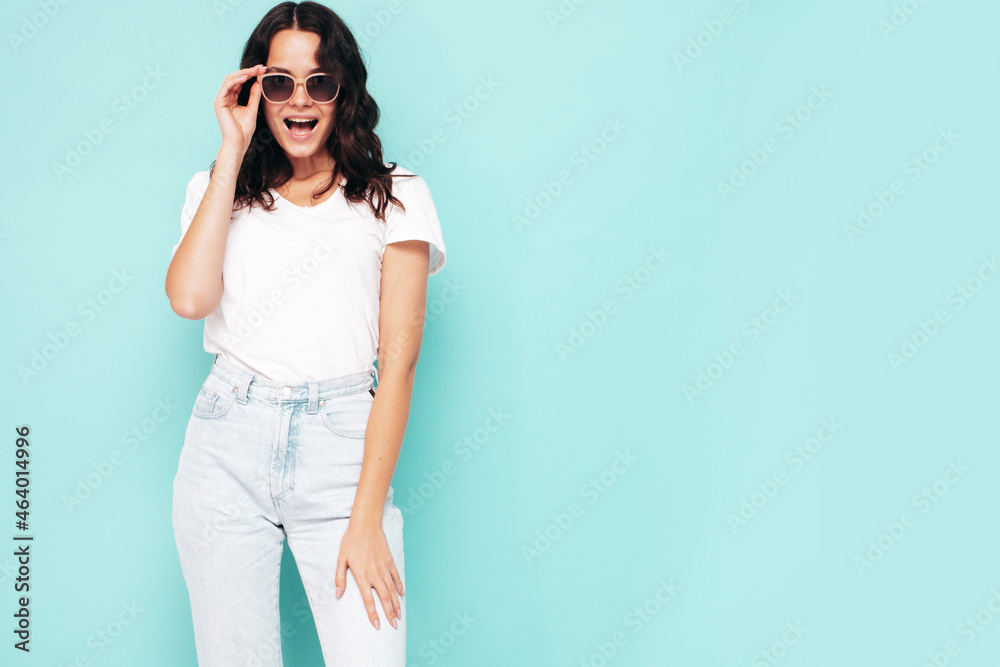 Portrait of young beautiful smiling female in trendy summer clothes. Sexy carefree woman posing near blue wall in studio. Positive model having fun indoors. Cheerful and happy. in sunglasses