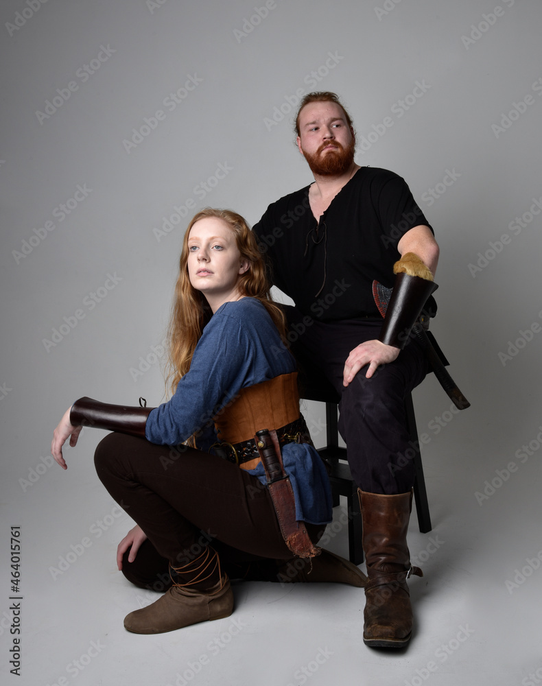 Fototapeta premium Full length portrait of red haired couple, man and woman wearing medieval viking inspired fantasy costumes, sitting poses, isolated on white studio background.