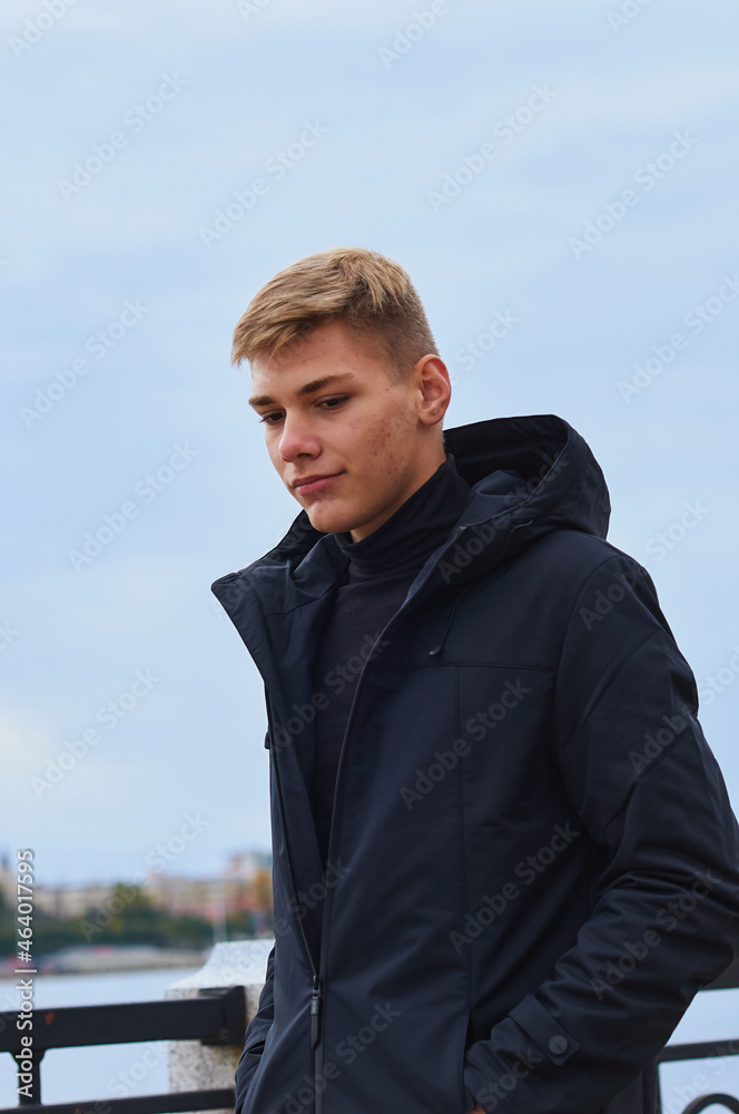 A handsome blond guy in a black hooded jacket walks along the river embankment. Look down. A handsome blond guy in a black hooded jacket walks along the river embankment. Look down.