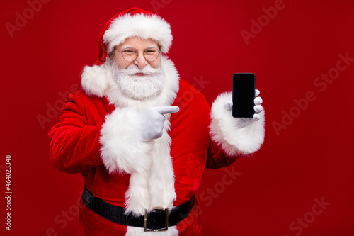 Photo of handsome funky man pensioner dressed santa claus costume pointing modern device empty space isolated red color background