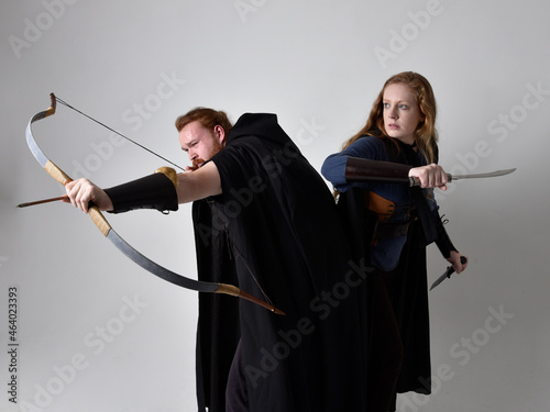 Full length  portrait of red haired  couple, man and woman wearing medieval viking inspired fantasy costumes, standing fighting pose holding  archery bow and arrow and long sword weapons, isolated on 