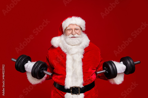 Photo of strong funky retired man wear santa claus costume spectacles holding heavy dumbbells isolated red color background