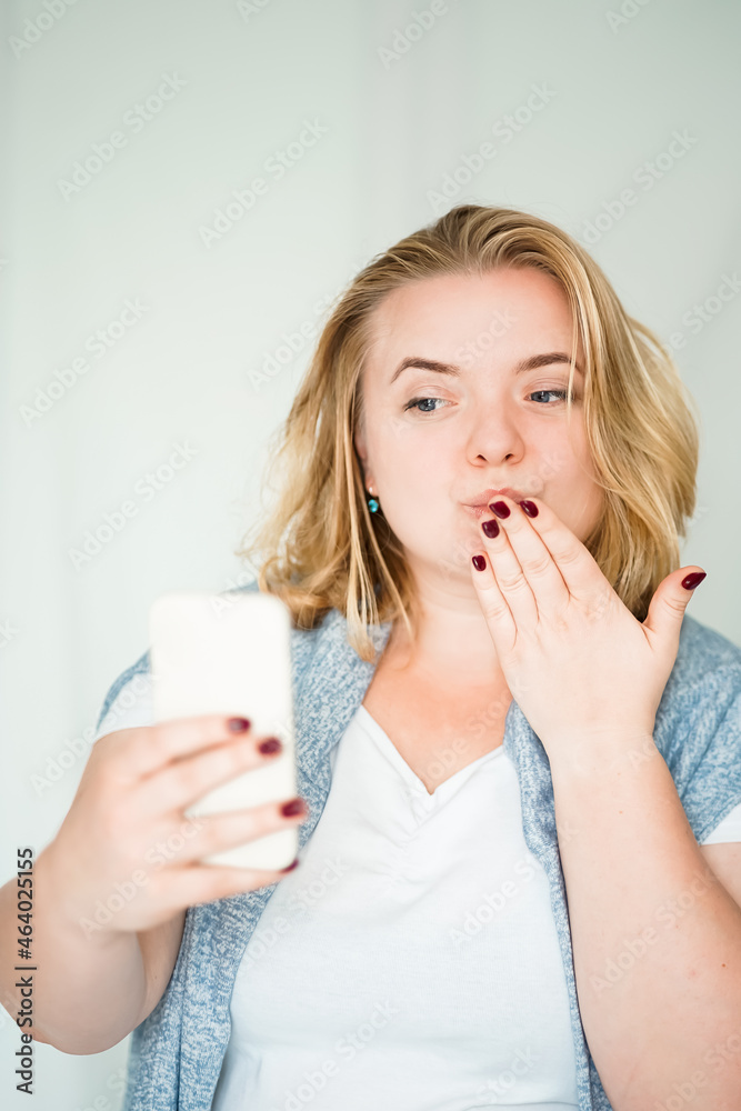 blonde woman sends air kiss to mobile phone on white wall background. human emotions.