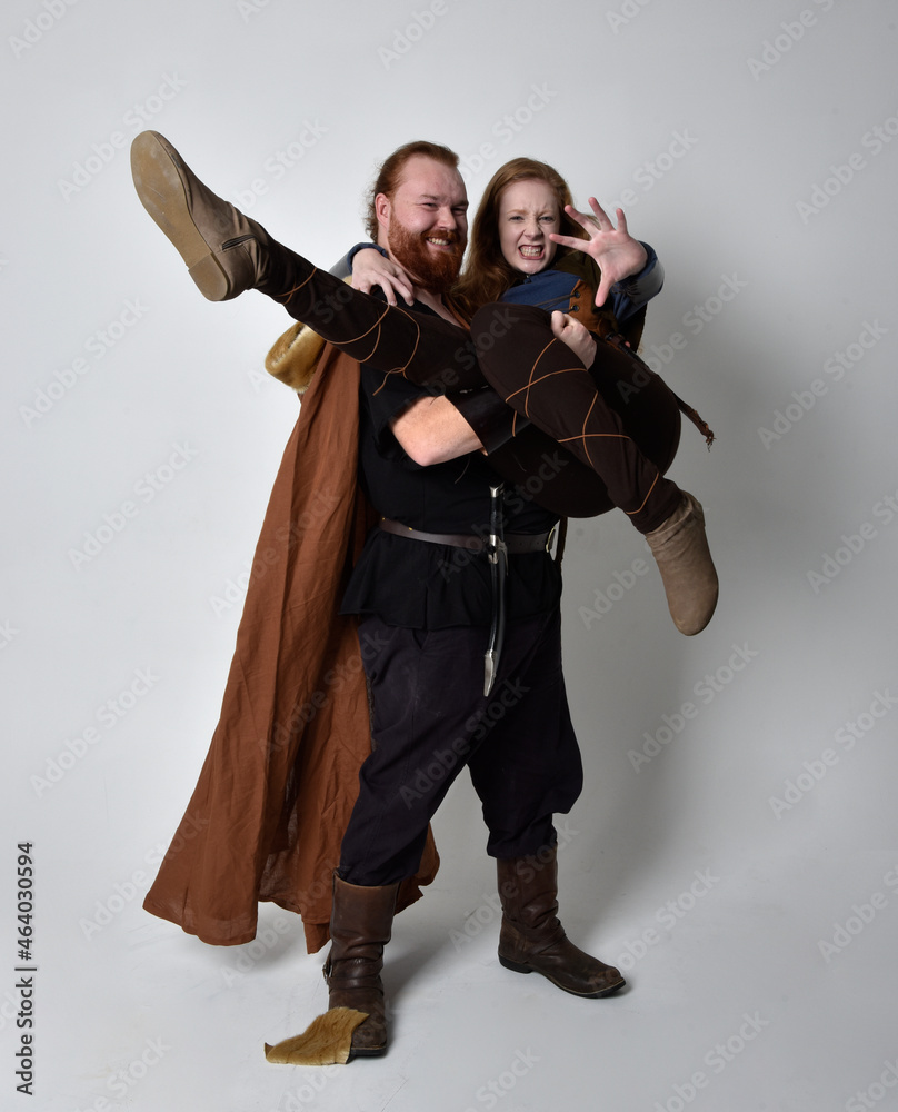 Fototapeta premium Full length portrait of red haired couple, man and woman wearing medieval viking inspired fantasy costumes, standing pose, isolated on white studio background. 
