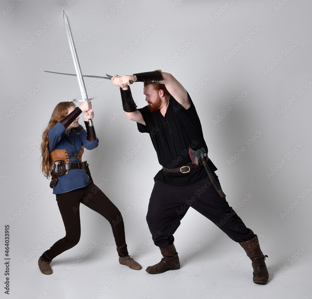 Full length portrait of red haired couple, man and woman wearing medieval viking inspired fantasy costumes, standing fighting pose holding sword weapons, isolated on white studio background. Full length portrait of red haired couple, man and woman wearing medieval viking inspired fantasy costumes, standing fighting pose holding sword weapons, isolated on white studio background.