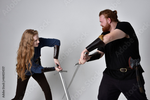 Full length portrait of red haired couple, man and woman wearing medieval viking inspired fantasy costumes, standing fighting pose holding sword weapons, isolated on white studio background.