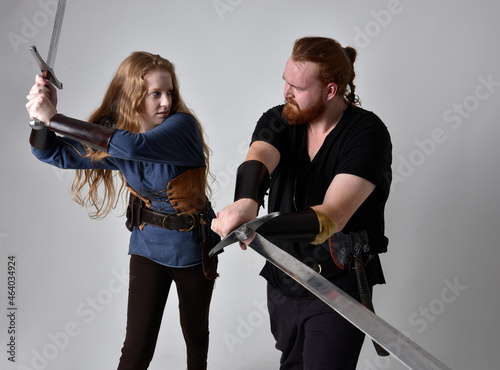 Full length portrait of red haired couple, man and woman wearing medieval viking inspired fantasy costumes, standing fighting pose holding sword weapons, isolated on white studio background.