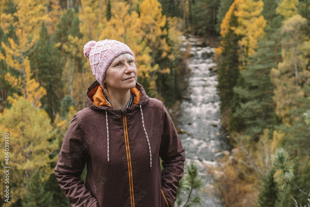An authentic portrait of an adult female traveler in warm clothes on top of a mountain, against the background of an autumn forest. Looks up. Eco tourism. Selective focus. Close-up.
