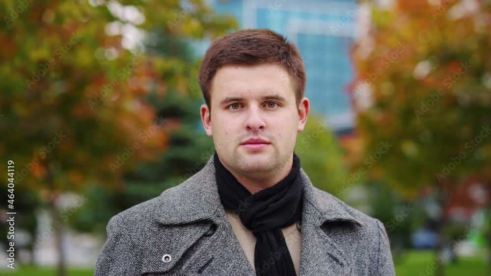 Young man with short haircut wearing coat and scarf posing in autumn park, green and orange trees on blurred background. Portrait of male model outside. Concept of season Young man with short haircut wearing coat and scarf posing in autumn park, green and orange trees on blurred background. Portrait of male model outside. Concept of season