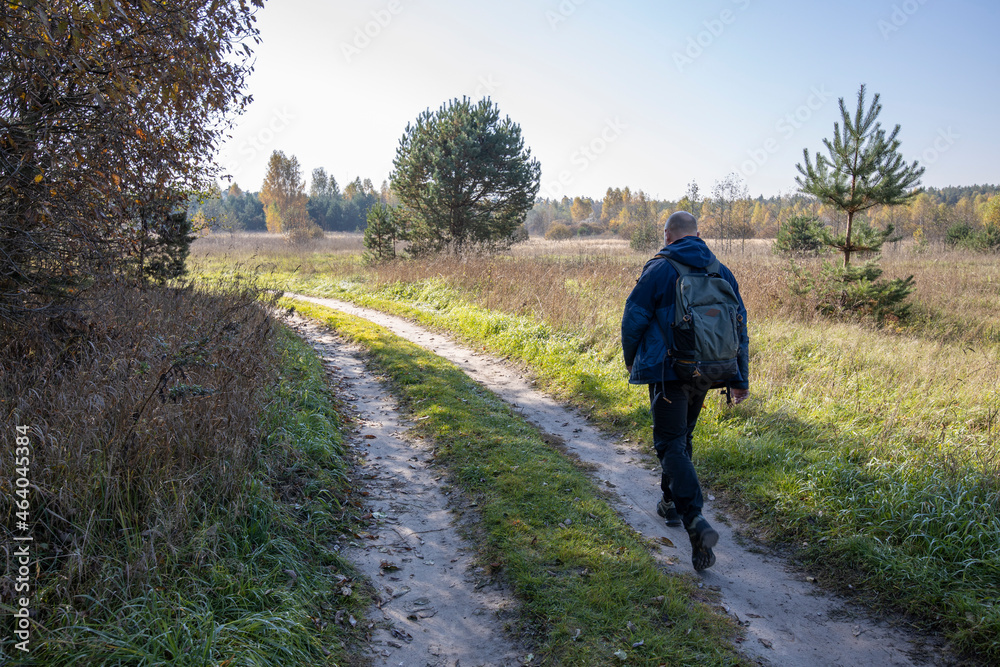 A tourist walks along a country road early in the morning. A man with a backpack on the background of a field illuminated by sunlight.