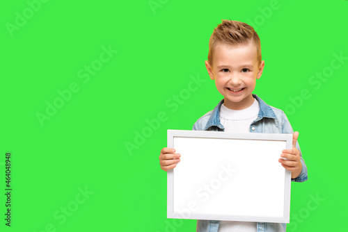 Five year-old boy smiles and holds blank white frame. Happy preschooler child isolated on green background with copy space for message, information, mock up. Kid hold a big photo frame.