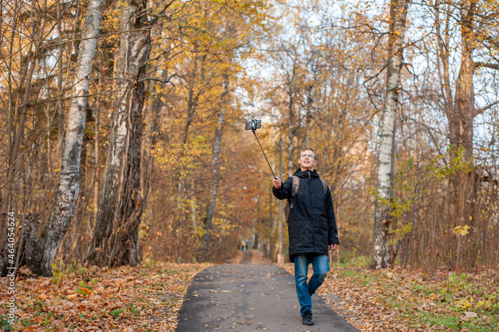 A teenage boy walks through an autumn park and shoots a video. A Caucasian boy in a black jacket holds a monopod with a smartphone. Blogger. Emotional communications.. A teenage boy walks through an autumn park and shoots a video. A Caucasian boy in a black jacket holds a monopod with a smartphone. Blogger. Emotional communications..