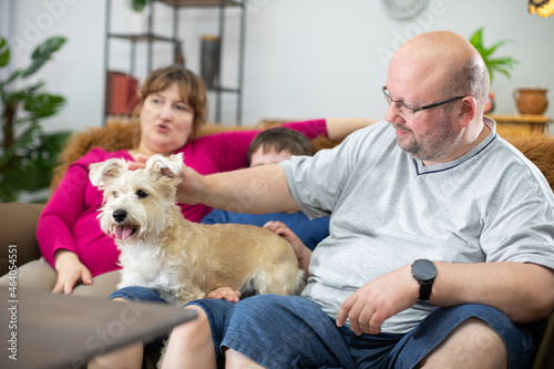 A father strokes a shaggy dog while sitting on the sofa with his family, his son and wife.