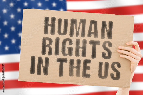 The phrase " Human rights in the US " on a banner in men's hand with blurred American flag on the background. Dictatorship. Equality. Freedom for people. Restrictions for civil citizens. Government © AndriiKoval