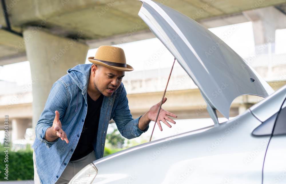 Frustrated young asian man looking at broken down car engine on street waiting for assistance to arrive and recover or fix the vehicle. Male angry stand front a broken car. Broken car while traveling. Frustrated young asian man looking at broken down car engine on street waiting for assistance to arrive and recover or fix the vehicle. Male angry stand front a broken car. Broken car while traveling.