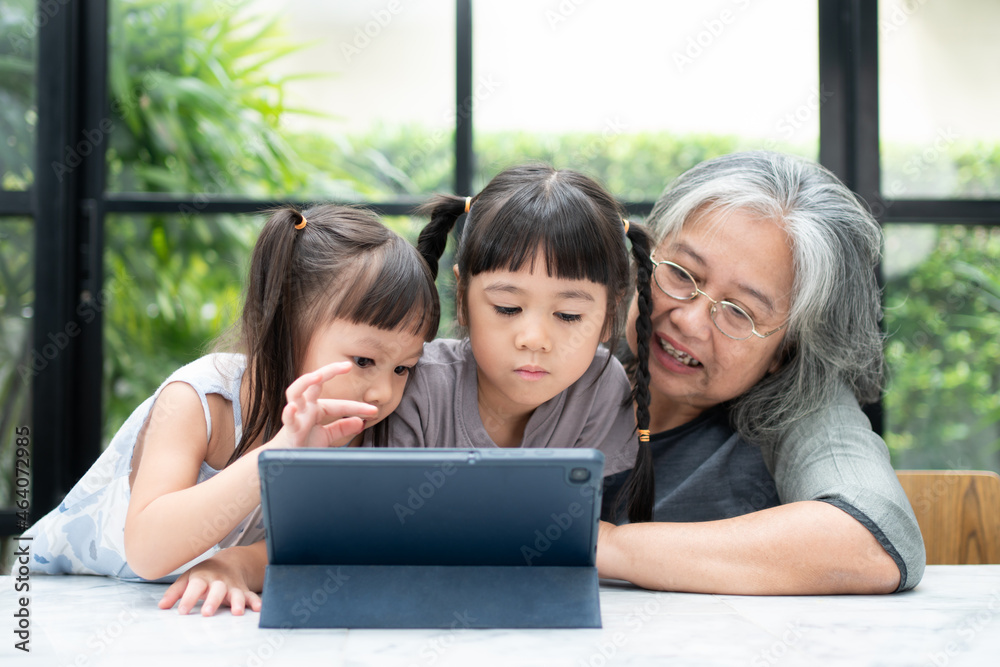 Asian Grandmother with her two grandchildren having fun and playing education games online with a digital tablet at home in the living room. Concept of online education and caring from parents.