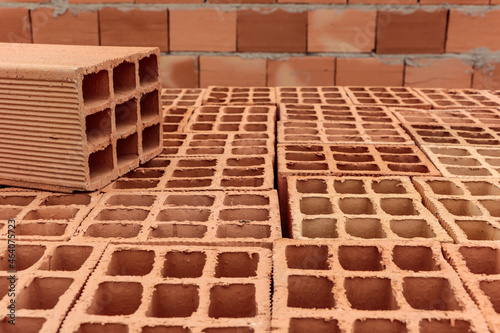 Pile of clay bricks used for building masonry house. In the image the bricks are laid out showing their holes  with one above them highlighted  and in the background an already built wall. 