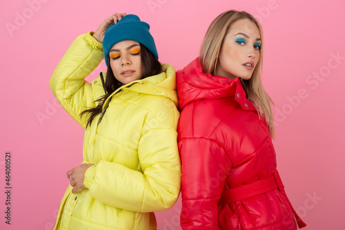 two attractive stylish women posing on pink background in colorful winter down jacket of red and yellow color, warm clothes fashion trend